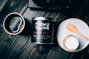 Embalagem do Geisha Passa do Moka preta com escritos em roxo. Ao lado, um copo de leite, um de café e uma colher de pau.